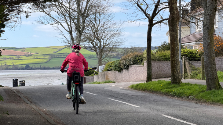 A person cycling on the road towards the coastline; water and green fields on a hill in the background.
