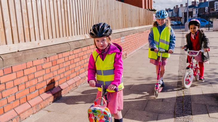 An image of school children riding scooters, wearing helmets, in an urban setting.