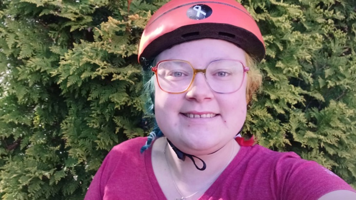 A young white woman smiling wearing a pink jumper in front of a hedge outside her home in Manchester