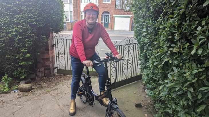 A young white woman smiling wearing a pink jumper stood with her electric foldable cycle outside of her home in Manchester