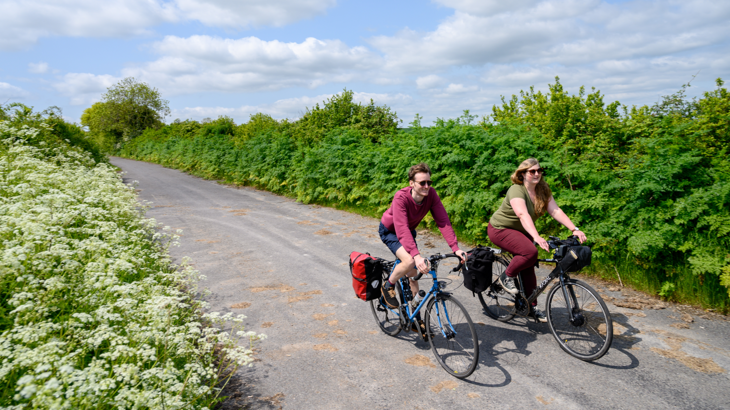 two people cycle happily on a newly repurposed road, now a traffic free walking wheeling and cycling route lined with greenery on a sunny day.