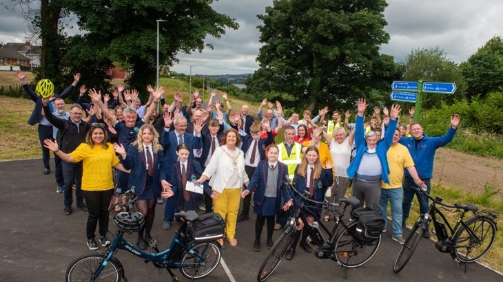 A large group of male and female of all ages are gathered with their arms in the air on a wide traffic-free path with a number of bicycles in front.