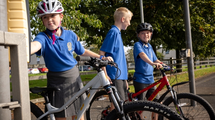 A schoolgirl on a bike wearing a helmet pushes the button on a pedestrian crossing while her two classmates stand and wait beside her.