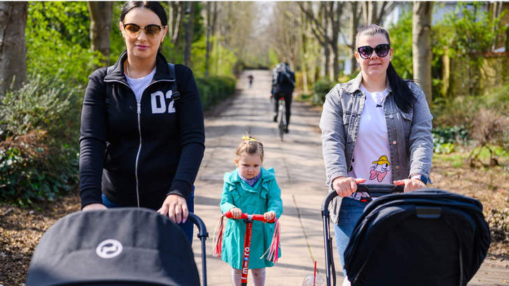 Two parents walking along a traffic-free route with pushchairs, and a little girl on her scooter in between them.