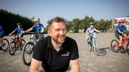A man wearing a black Walk Wheel Cycle Trust top smiles at something off camera with children on bikes behind him in a school playground.