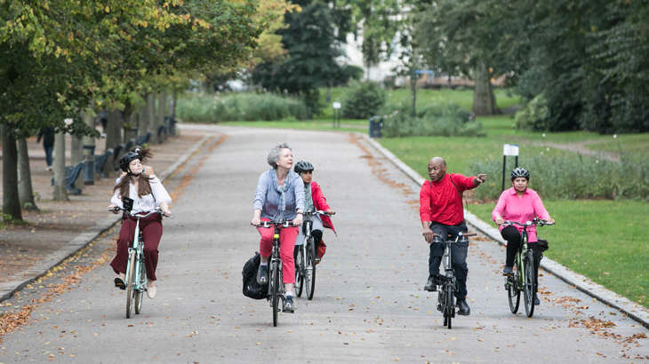 A group of people smiling as they cycle through a park in London, surrounded by trees.