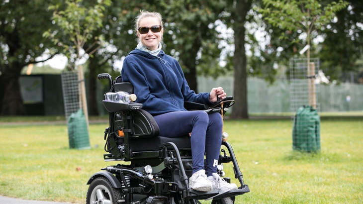 A person in a motorised wheelchair smiling as they wheel through a park.