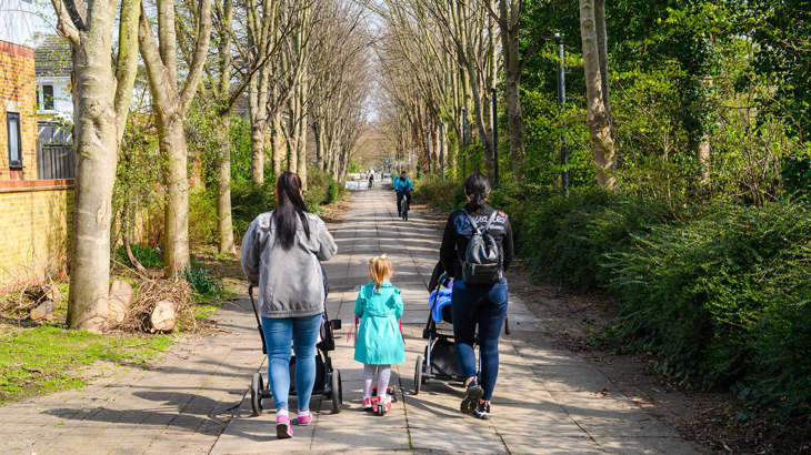 Two mums with pushchairs and a little girl on a scooter walking away from the camera along a traffic-free route surrounded by trees.