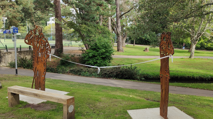 Two steel portraits beside a wooden bench. A ribbon is tied between them. A path runs behind. A field, lined with trees, is in the background.