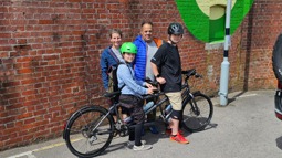 Paul Luttrell stands with his wife and two youngest sons as they sit on a tandem bike together in the sunshine.