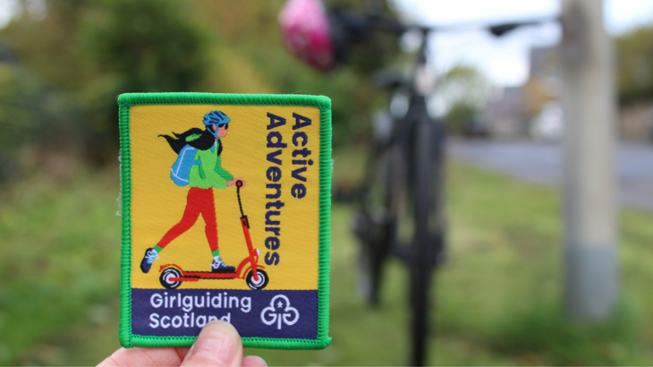 A colourful fabric badge with a cartoon of a girl on a scooter on it with a bike and a helmet in the background which are out of focus.