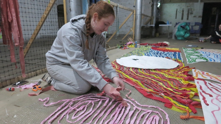A young artist kneels on the floor and works with materials to create an artwork.