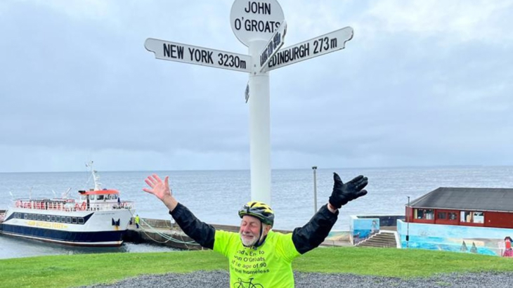 An older man on his knees with his arms in the air looking relieved at the Land's End signpost at the end of a long cycle across the length of the UK