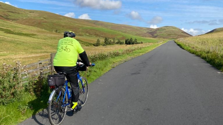 An older man with a high vis top on cycling up a hill on a tarmac road in the countryside with blue skies ahead
