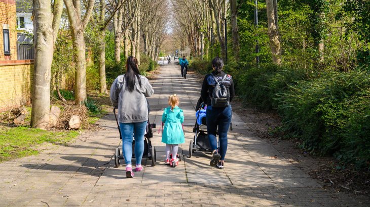 Two Women And A Young Girl Walking With Prams along a tree-lined path with a cyclist in the background