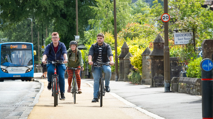 Three young adults cycling along a cycle path next to a road which has a 20mph sign and a bus on it