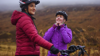 Two women wearing waterproof coats and cycling helmets on National Route 7 in the Scottish Highlands.