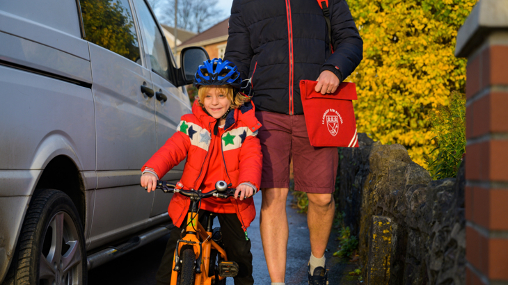 A boy on a bike with a helmet on in the sun navigating around a van which is parked on the pavement with an adult who's holding his primary school bag