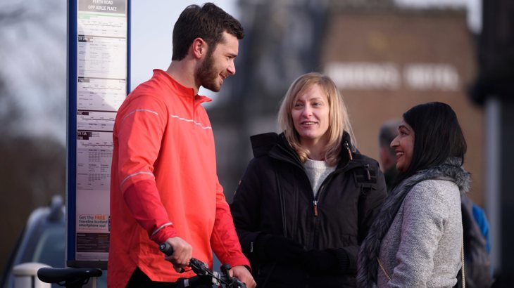 Three young people standing at a bus stop talking, one with a bike