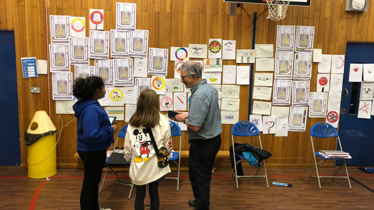 Two primary school pupils stood with an older man facing a wall full of leaflets and drawings in their sports hall