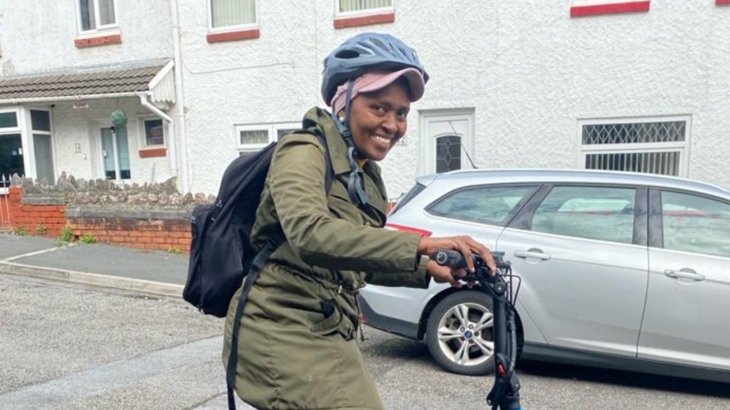 An E-Move project beneficiary smiling whilst looking at the camera, stood astride her e-bike on a residential street.