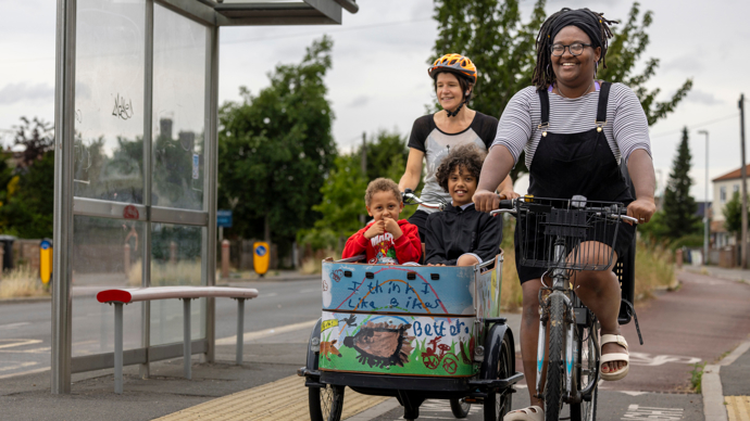 Two people smiling while cycling in a cycle lane in an urban area, one is cycling a cargo bike with two small, smiling children sat in it
