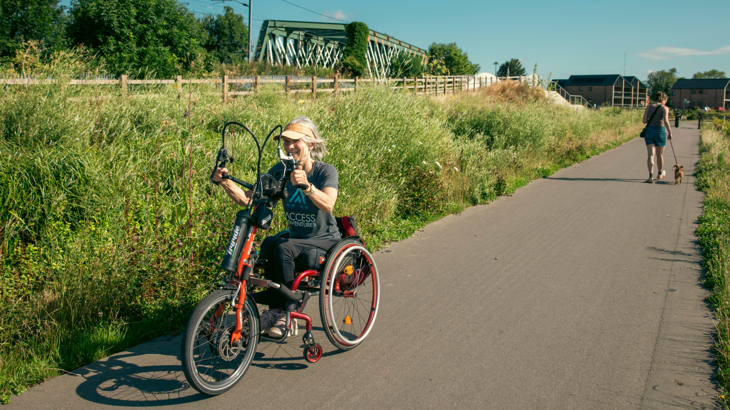A smiling woman using an orange hand bike on a path surrounded by greenery on a sunny day in the background a woman walks a small dog on the lead