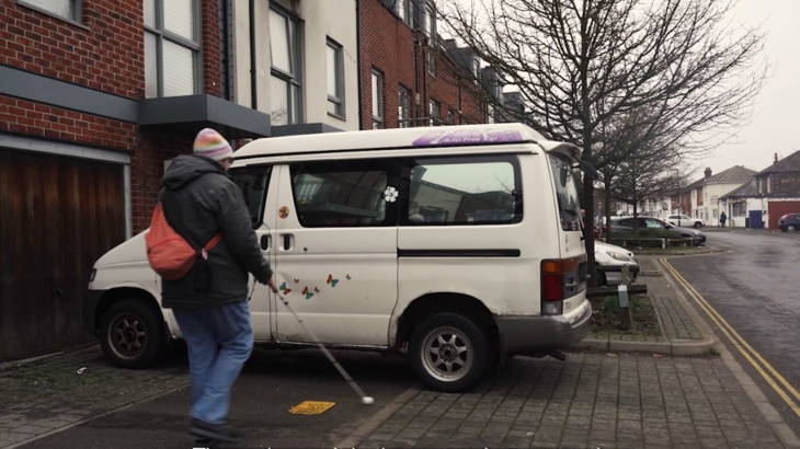 Person walking with a white cane facing a barrier in front of them, as a white van is parked on the pavement with the road next to them on the right.