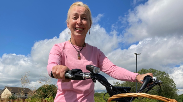 A woman sat smiling on her trike holding onto the handlebars wearing a pink long-sleeved top on a sunny day with blue skies in the background