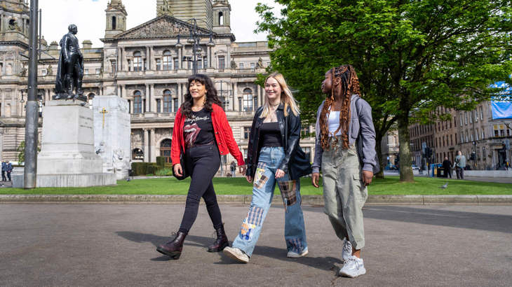 Three young women are walking through George Square in Glasgow City Centre, with City Chambers and a statue visible in the background.