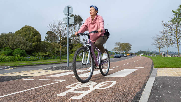 A lady dressed in pink cycles along a segregated cycle route in Broughty Ferry, with trees and the shoreline visible in the background.