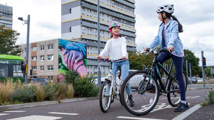 Two young females pause along a segregated cycling route in Woodside, Glasgow. A brightly coloured mural and towerblock in the background.