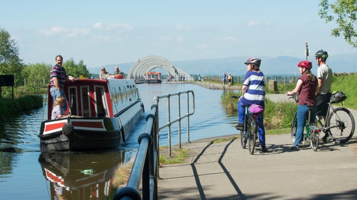 People walking and cycling along a canal with a narrowboat on the water.
