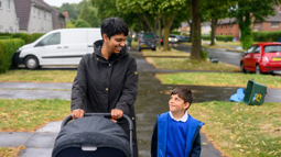 A parent pushing a pram in a residential area on a rainy day looking down at her child who is wearing a blue uniform and smiling looking up at her