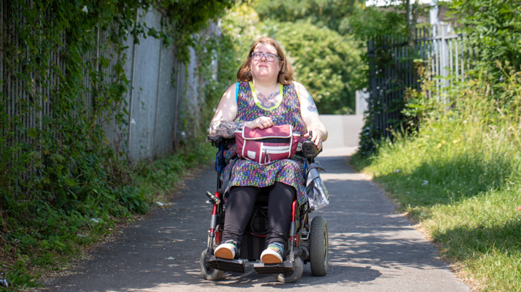 A person with tattoos and glasses in a powered wheelchair on an off-road cycle path in Liverpool on a sunny day surrounded by greenery