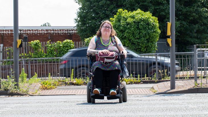 A person with tattoos and glasses in a powered wheelchair crossing a road at a Toucan crossing on a sunny day in Liverpool