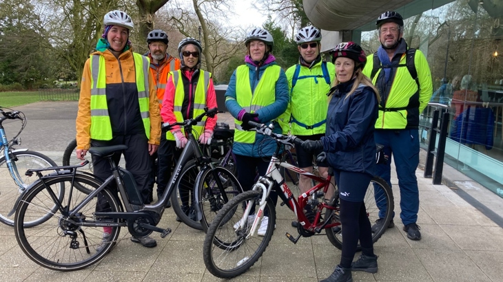 A group of men and women cyclists wearing helmets and hi-visibility vests pose for a photograph outside the Ulster Museum with Botanic Gardens behind.