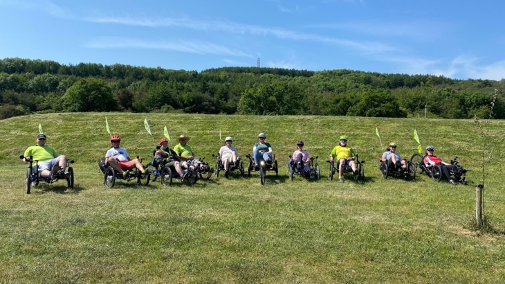 A group of recumbent nine trike users parked side-by-side on a grassy verge for a group photo, on a sunny day.