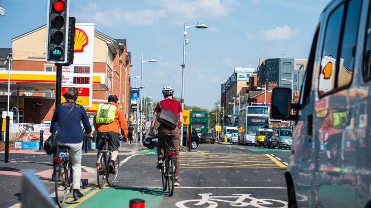 Three cyclists waiting a red traffic light alongside traffic. A petrol station is in the background.