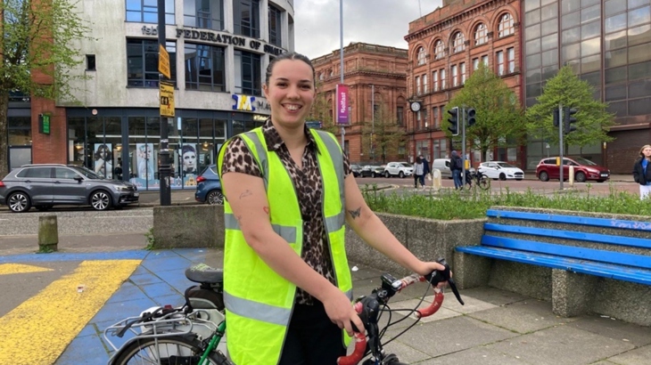 A woman wearing a hi-vis vest stands over a bike in Belfast city centre.