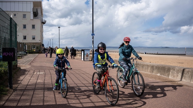 Two children and an adult are shown riding their bicycles on a traffic-free path next to a beach