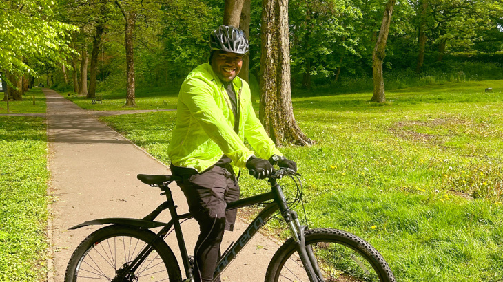 A man stood with his black cycle wearing high vis and a helmet smiling with grass and trees in the background on a sunny Spring day on a path