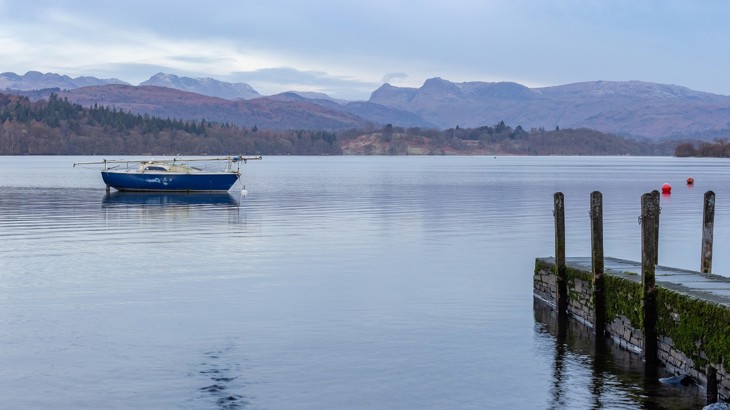 A pier on the shore of Lake Windermere, in a calm scene depicting a boat on the water and the mountains and foothills in the far off distance.