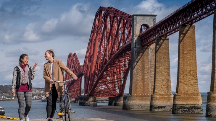 Two people walking along, one wheeling a bicycle, laughing and smiling as they chat while passing the Forth Bridge in the background.