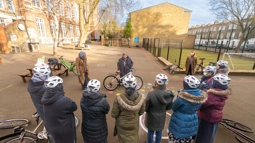 A group of mothers standing beside bicycles take part in a cycling workshop led by instructor. They are in the grounds of a school.