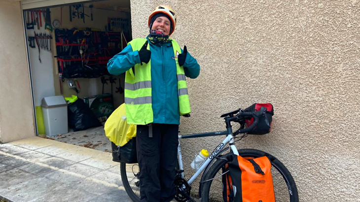 A woman stood smiling outside of a home with two thumbs up next to her cycle wearing a high vis vest, a blue ran jacket and a helmet