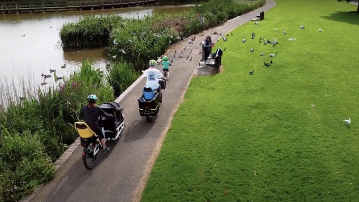 A family is shown from above riding cargo bikes through a park. A number of ducks are shown sitting on the path beside them.