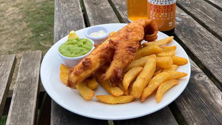 A plate of fish and chips with mushy peas on a wooden bench outside next to a bottle of beer