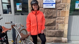 A woman wearing an orange jacket and black leggings and helmet stands beside bikes out an Active Travel Centre.