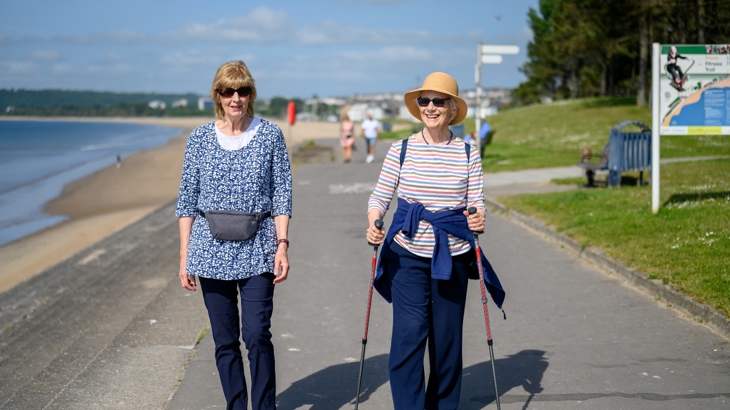 Two elderly women walking along Swansea Bay on the National Cycle Network on a sunny day.